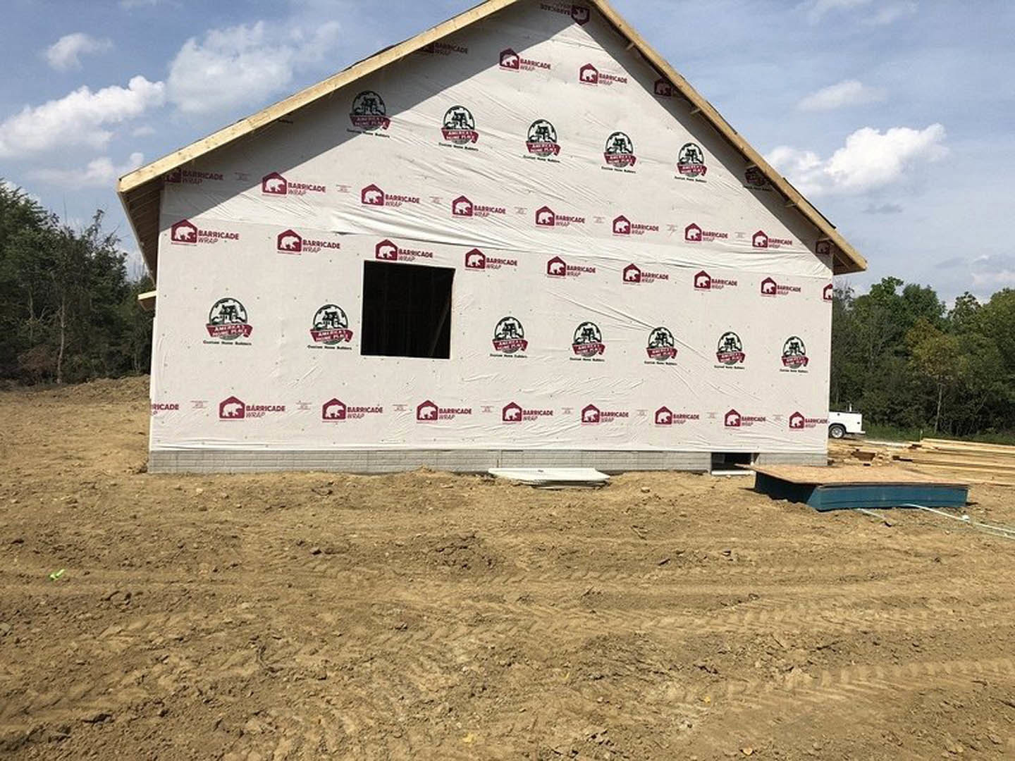 Partially built house wrapped in protective plastic sheeting, dirt field in foreground, white sign with red text near construction area, white wall with logo visible behind site