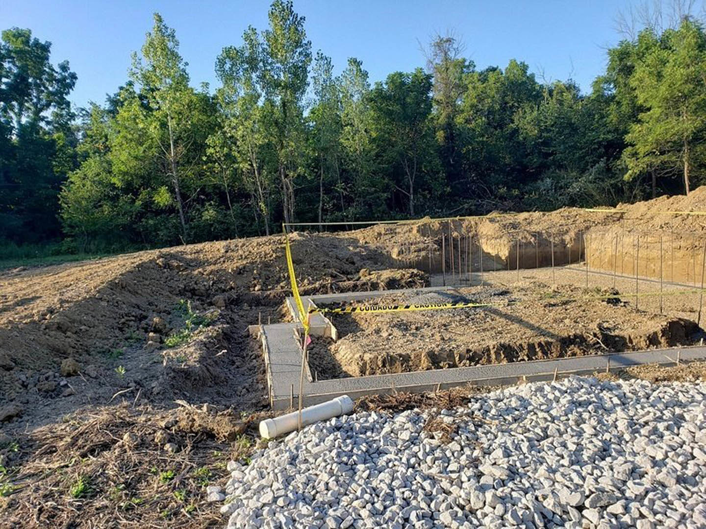 Dirt construction site bordered by yellow caution tape, scattered rocks and rubble, grassy patches, and a backdrop of tall trees under a clear sky