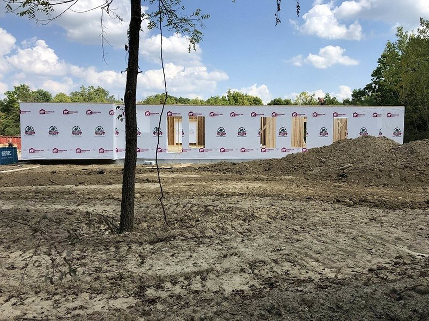 Framed custom home under construction with exposed wood, dirt lot, scattered trees, and blue sky with clouds