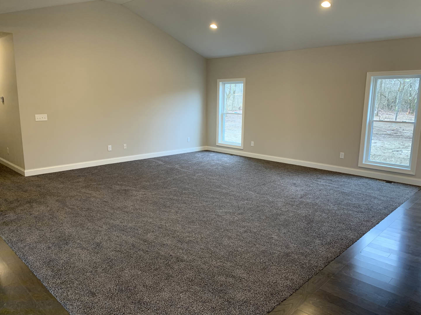 Spacious carpeted living area with large windows overlooking snowy forest, adjacent to wood flooring and white plaster walls, under a smooth ceiling.