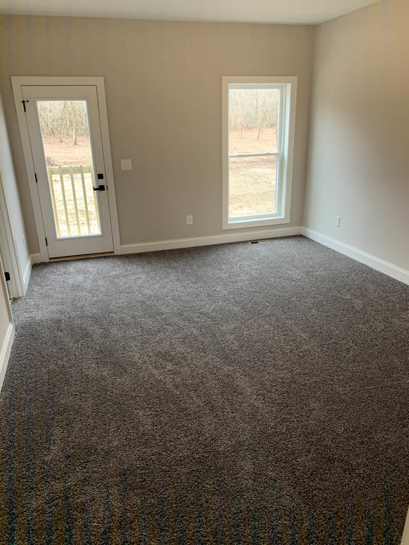 Carpeted room with white walls, single door, and large windows overlooking a dirt field, fence, and trees