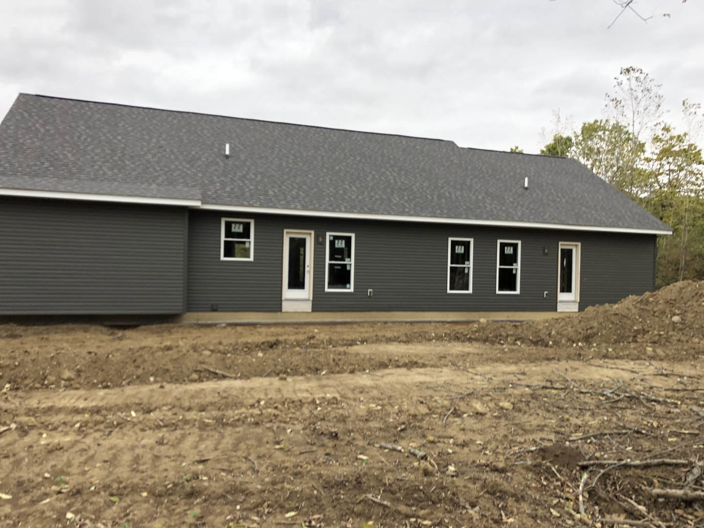 Grey house under construction with white door, black trim, white-framed windows, dirt yard, and surrounding trees
