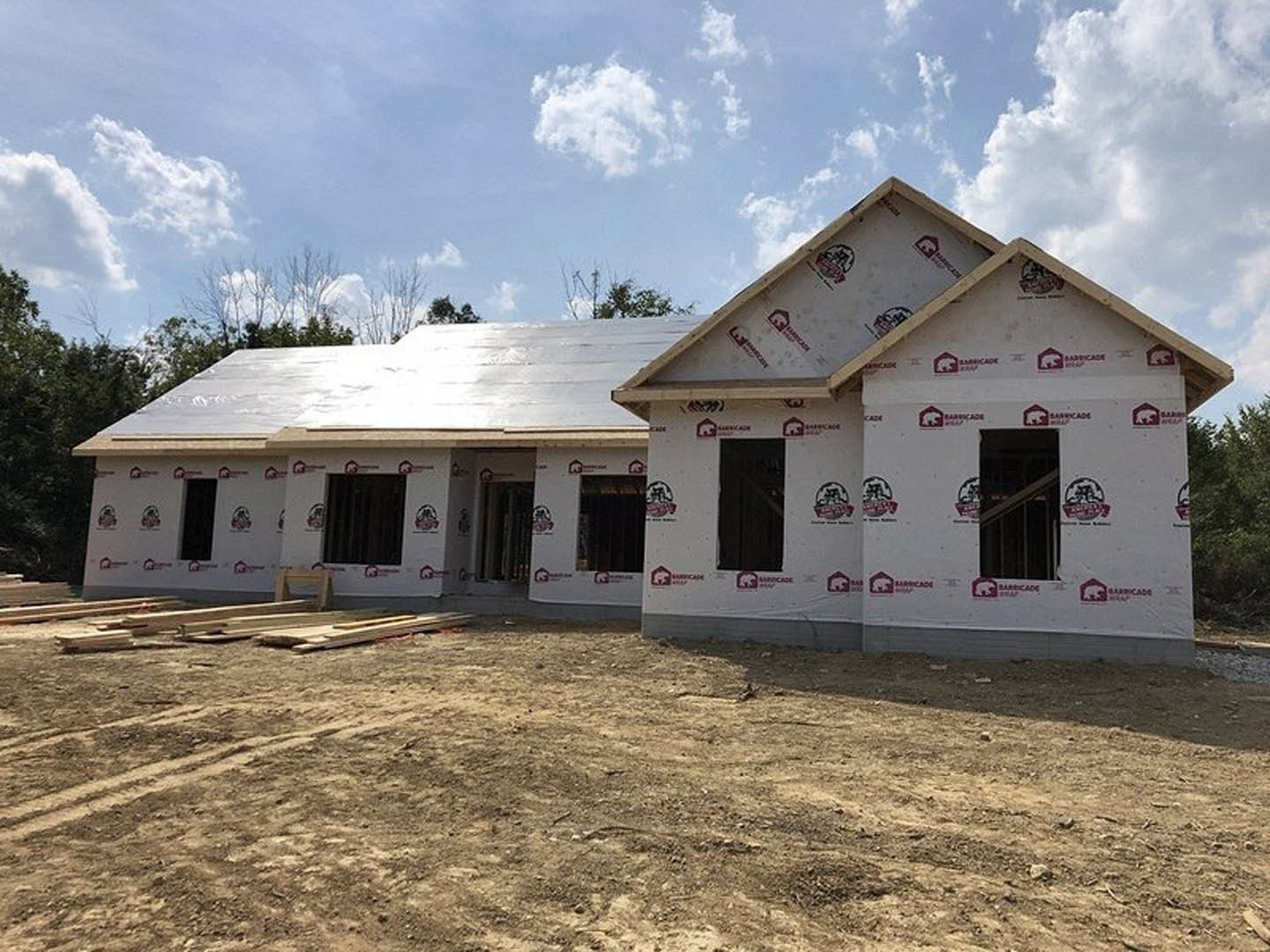 Two-story house under construction with exposed framing and roof, surrounded by dirt field and scattered white boards, cloudy sky overhead