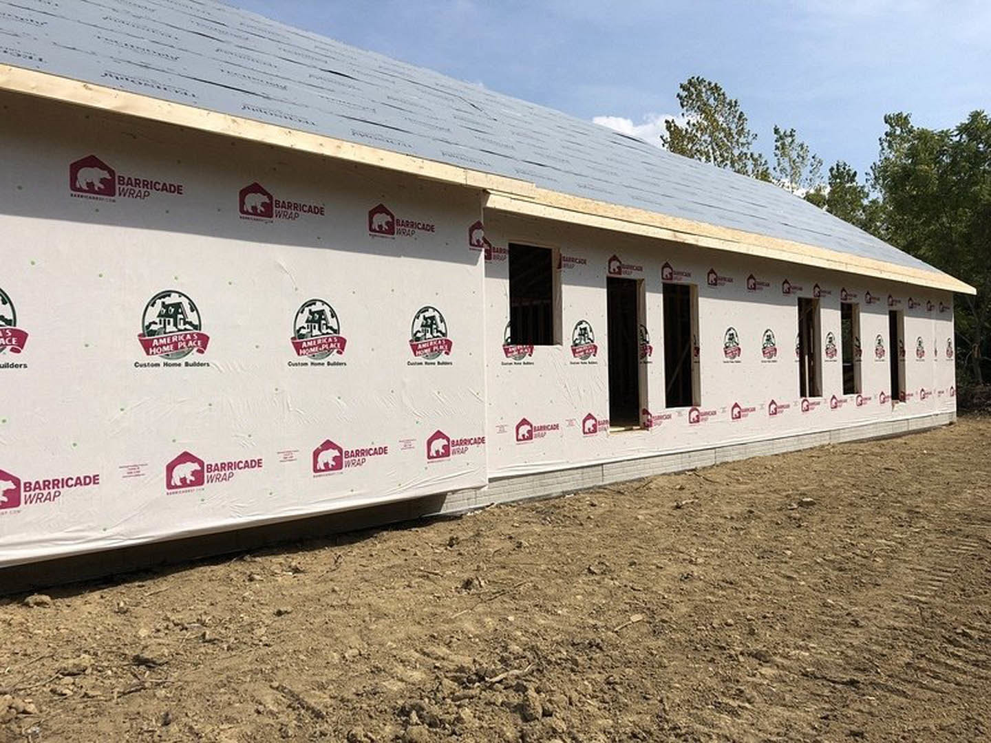 Partially built house with exposed framing, several installed windows, and white construction sheets featuring a red bear logo, surrounded by a dirt field under a cloudy sky.