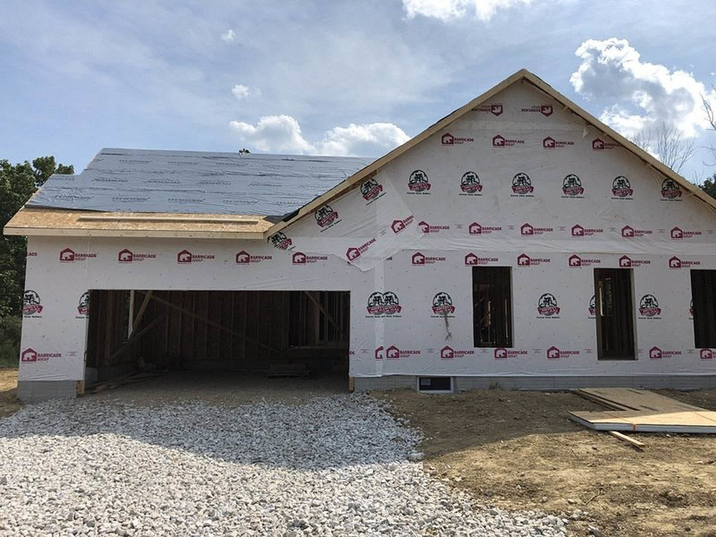 Partially finished house with shingled roof, exposed framing, and construction materials on dirt lot; cloudy sky and trees in background