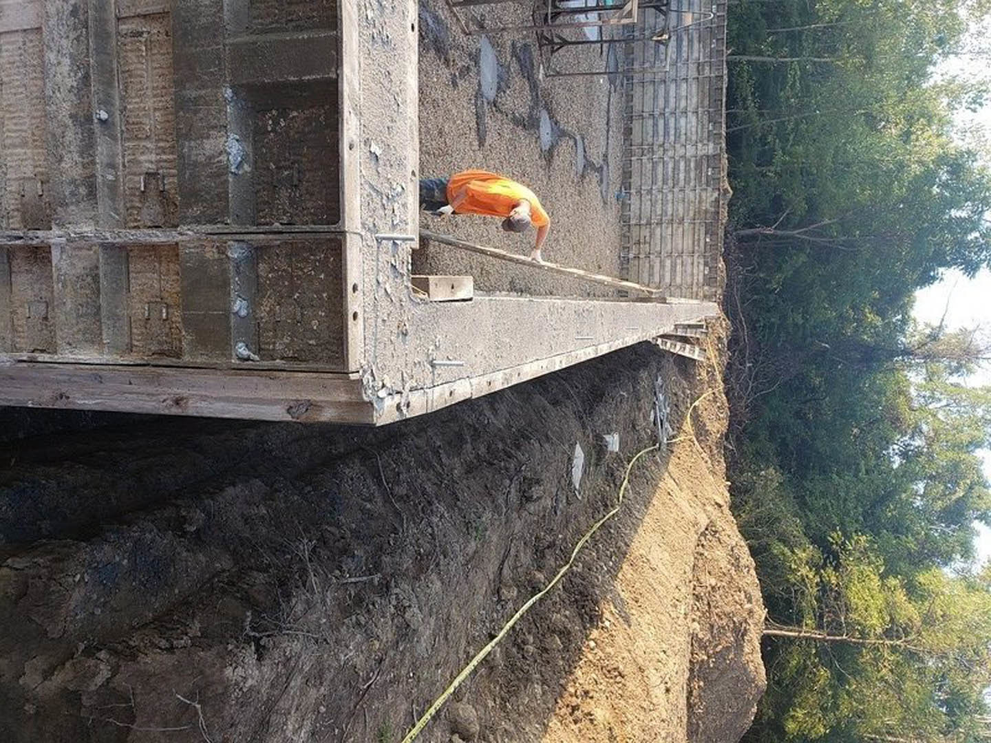 Man in orange shirt and blue jeans working on unfinished concrete structure outdoors, surrounded by trees and dirt path