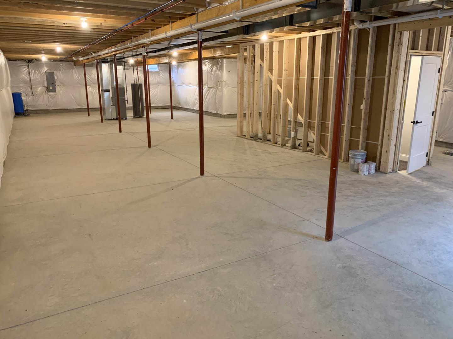 Basement room with exposed wood ceiling beams, visible metal pipes, concrete floor, white door with black handles, wooden shelving unit, ceiling pipe, and plastic storage container