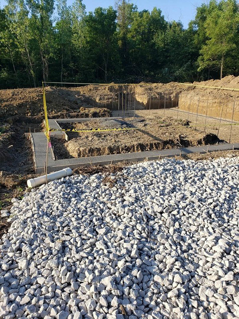 Gravel and rocks scattered across a construction site with exposed pipe, piles of dirt, metal rods, and surrounding trees under an open sky.