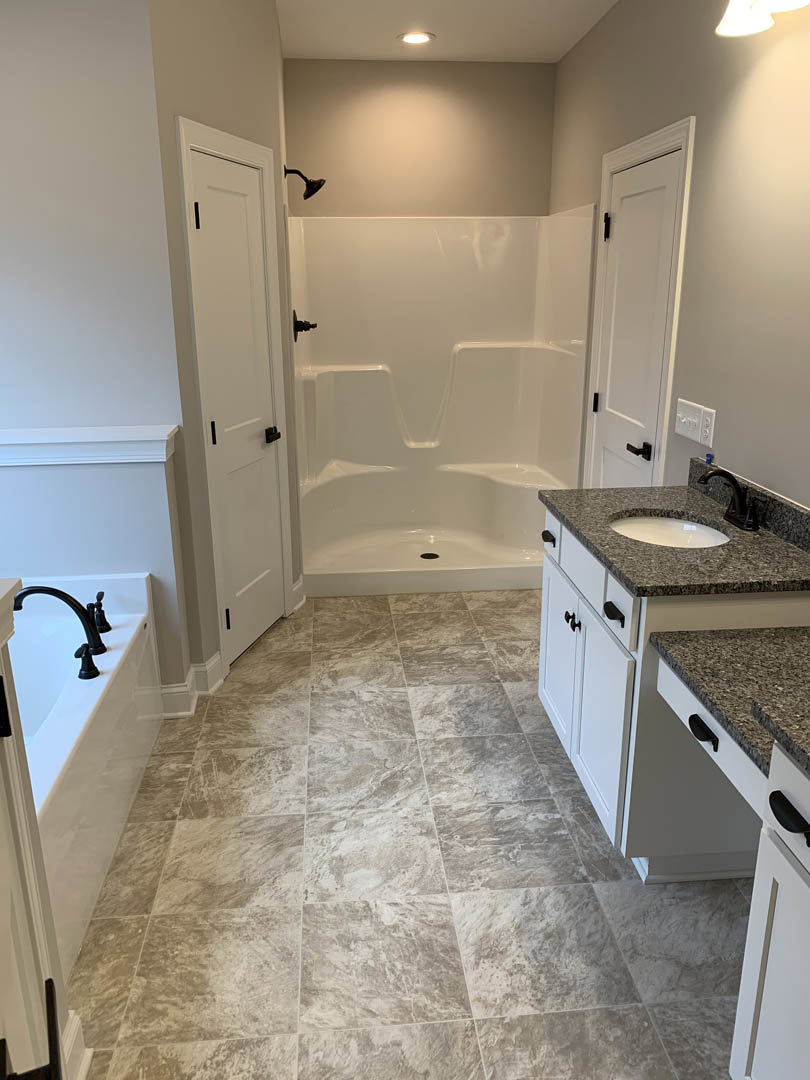 Bathroom with tile floor, freestanding tub, glass shower enclosure with black trim, white door featuring black handles, window showing blue sky, white walls, and silver hardware.