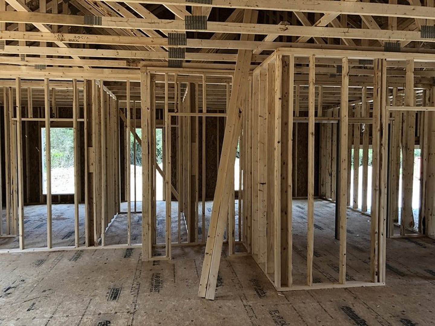 Exposed wooden beams and framing inside a house under construction, unfinished floors, open window showing outdoor view, floor vent visible