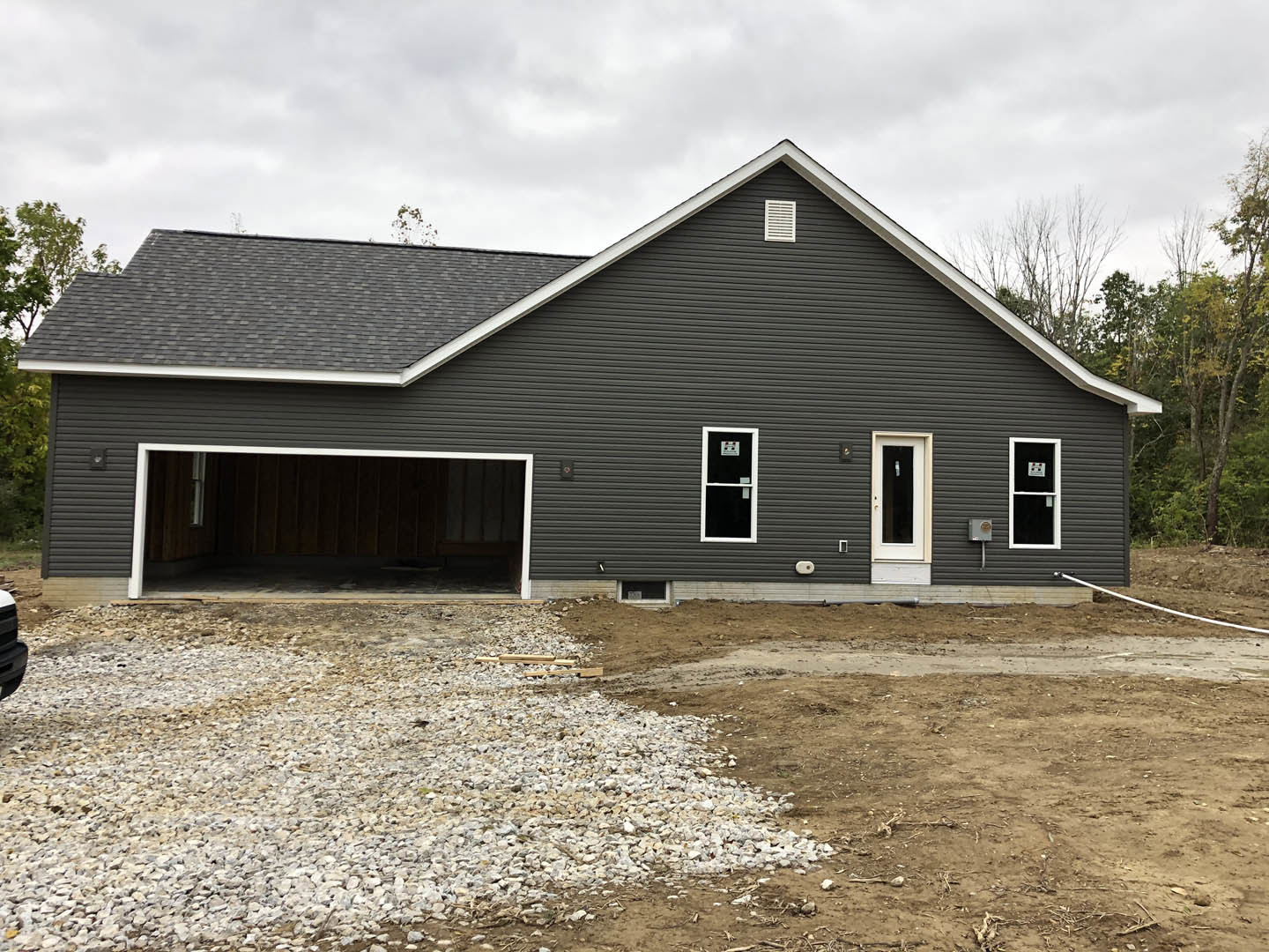 Two-story house with gray siding, attached garage, white framed door with window, person standing inside garage, pile of gravel and rocks in driveway, trees and cloudy sky in