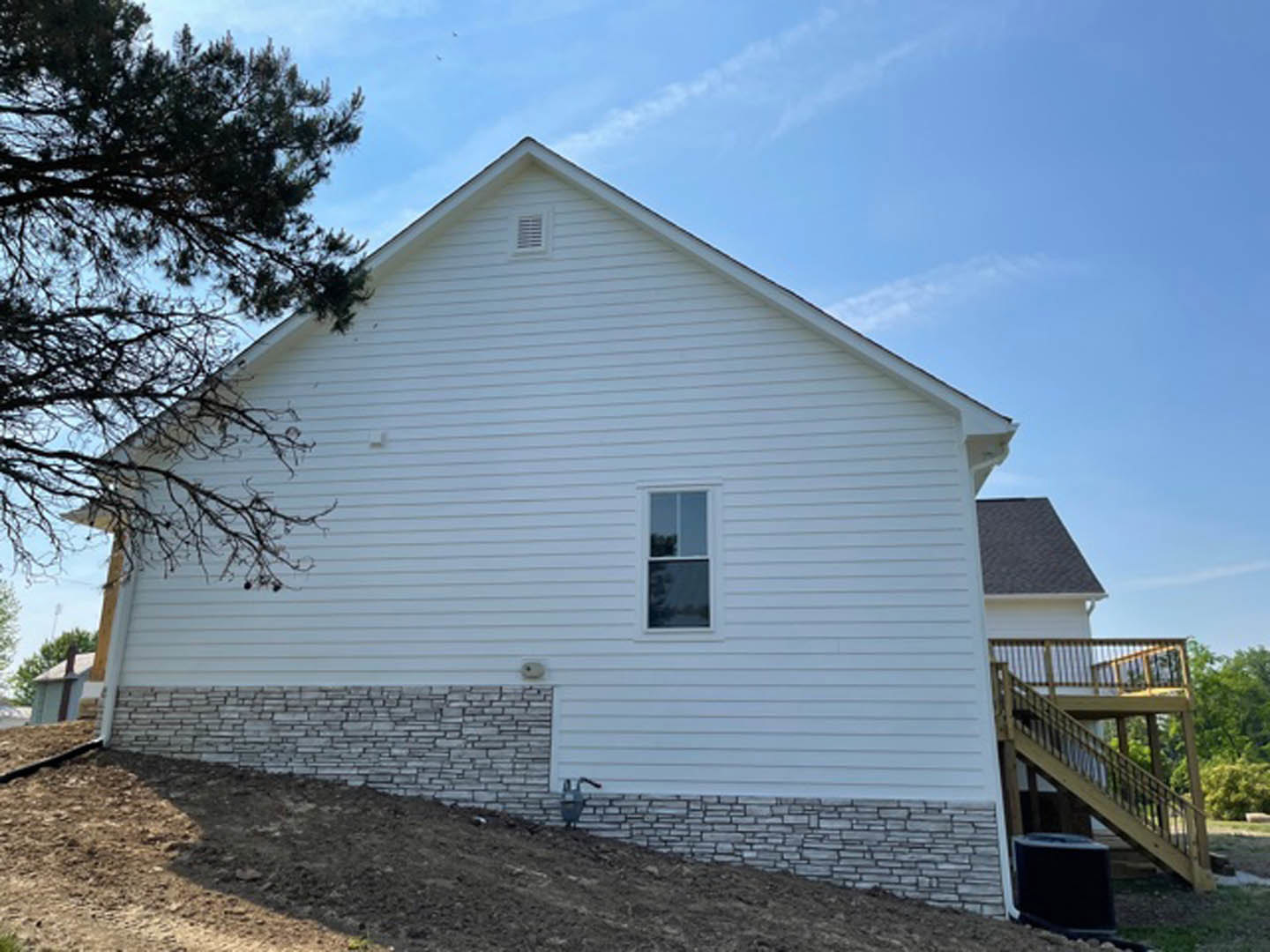 White cottage-style home with horizontal siding, wooden deck featuring railing, large tree beside house, white-framed window, exterior wall vent, partly cloudy sky above