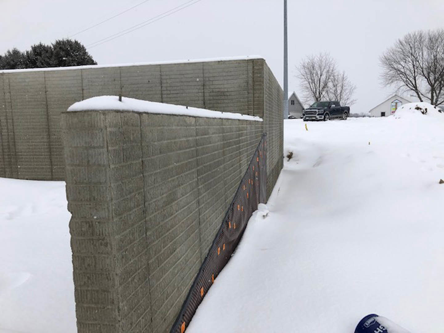 Snow-covered stone wall beside modern residential building, black car parked on plowed driveway, leafless tree and group of trees in snowy yard, overcast winter sky.