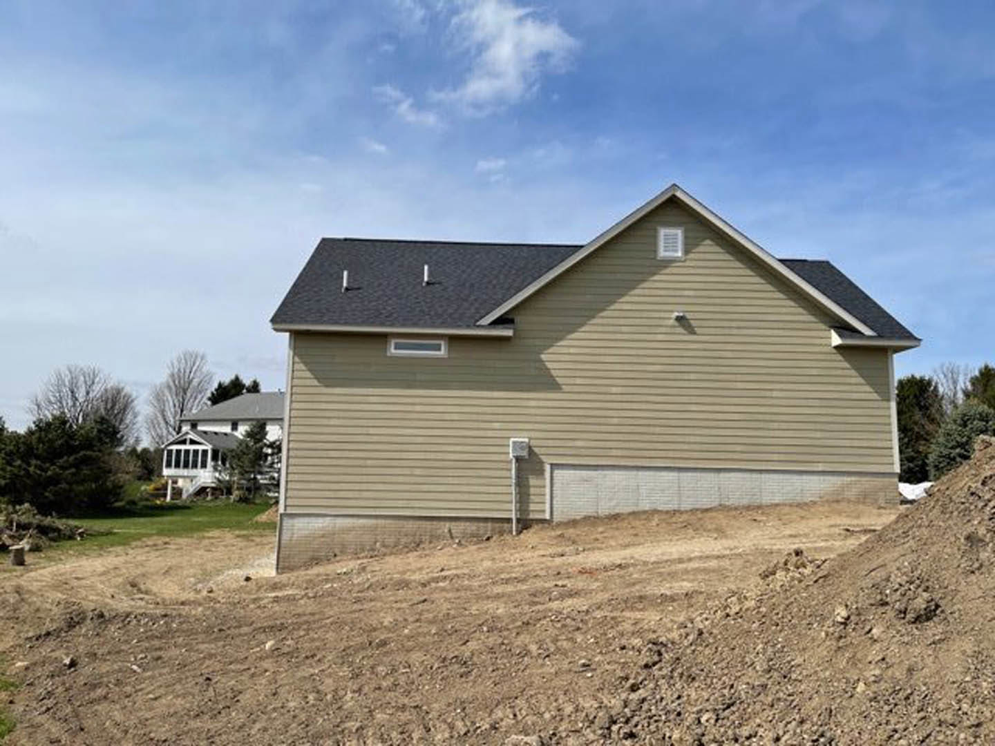 White brick house with brown roof and attached garage, situated on a dirt hill, front porch visible, surrounded by trees under a partly cloudy sky