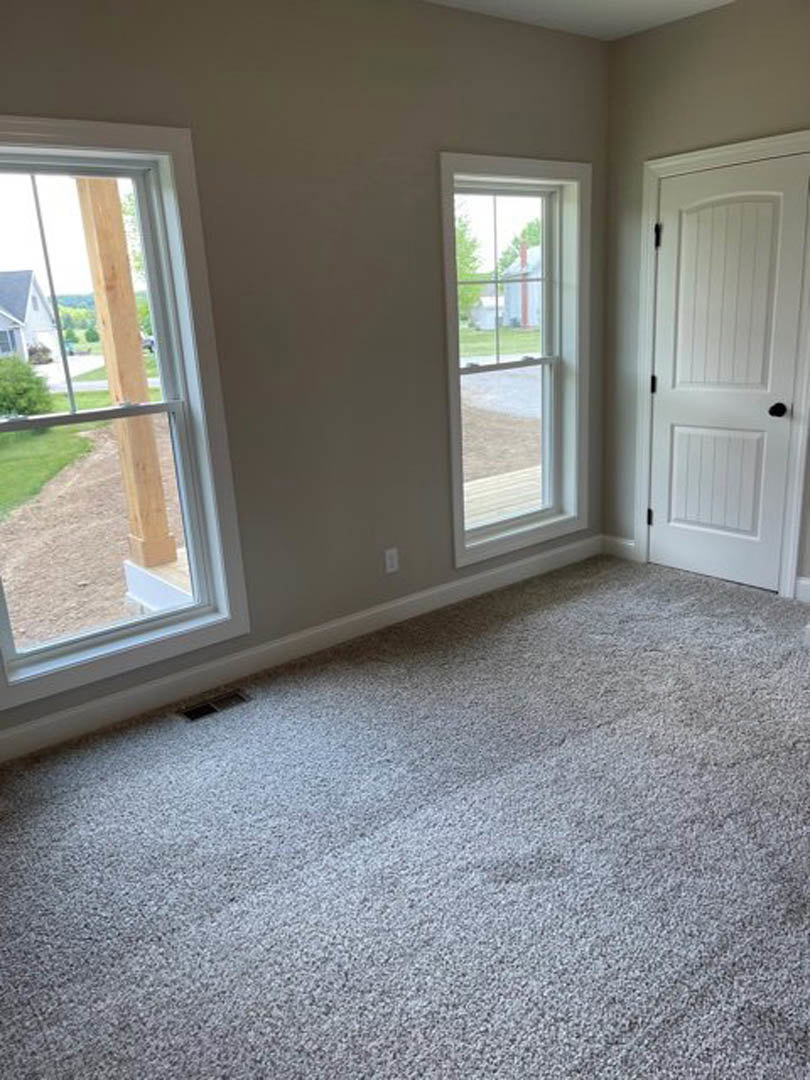 Bright room with white walls, white door featuring a black knob, two windows with wood beam accents, neutral carpet flooring, and outdoor greenery visible through the glass.