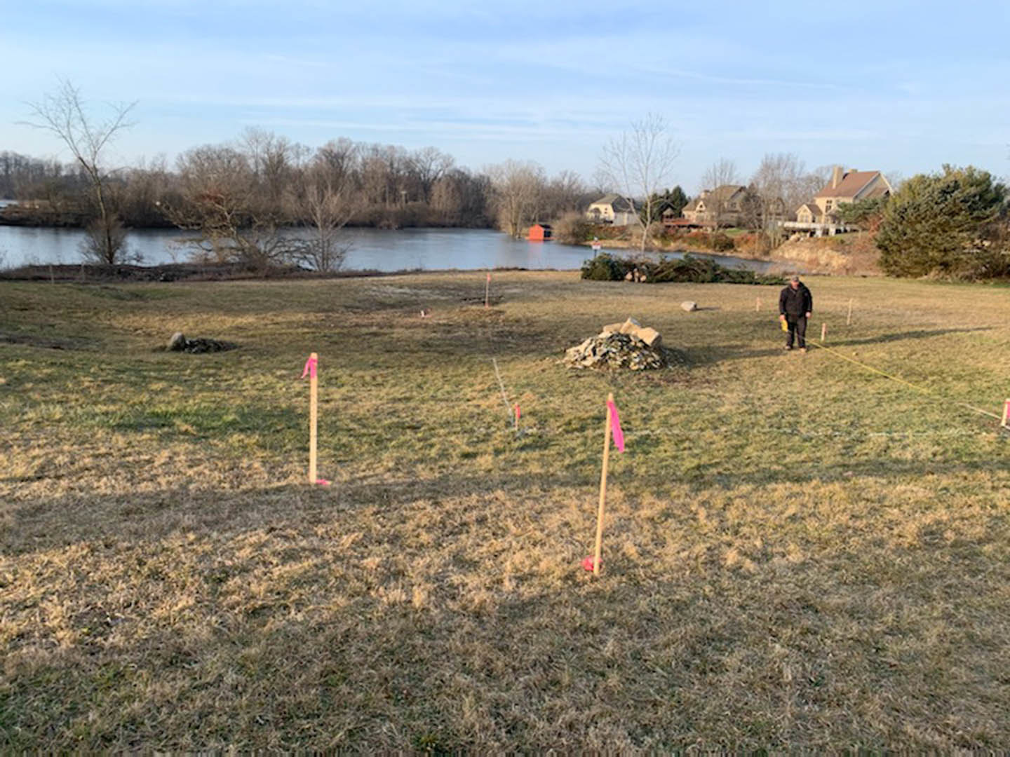 Modern home with stone chimney and large windows, surrounded by grassy field, scattered rocks, and mature trees near a lake under partly cloudy sky