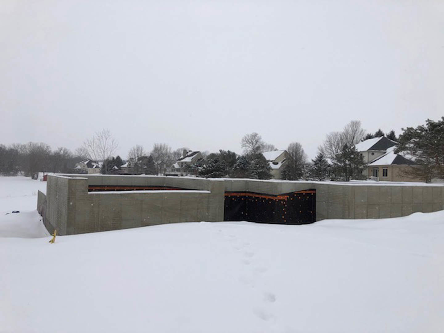 Modern house with black and orange facade, snow-covered ground with footprints, black gate leading through a tunnel-like entry, surrounded by trees and neighboring homes in a