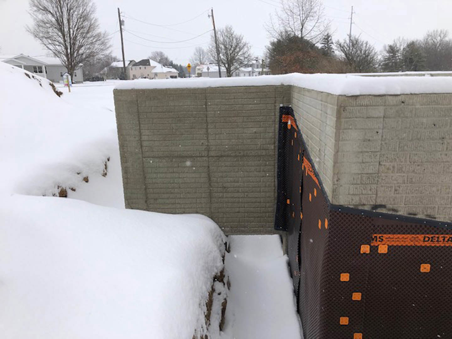 Snow piled against a textured concrete wall, bare tree branches and power lines visible in the wintry outdoor scene