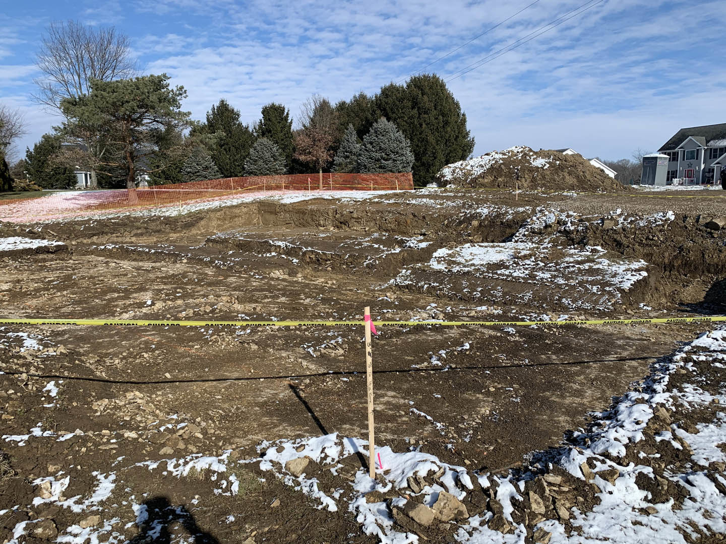 Dirt field with patches of snow, yellow caution tape, white post with black writing, large shed beside a house, fence dividing the field, group of trees and power line in the