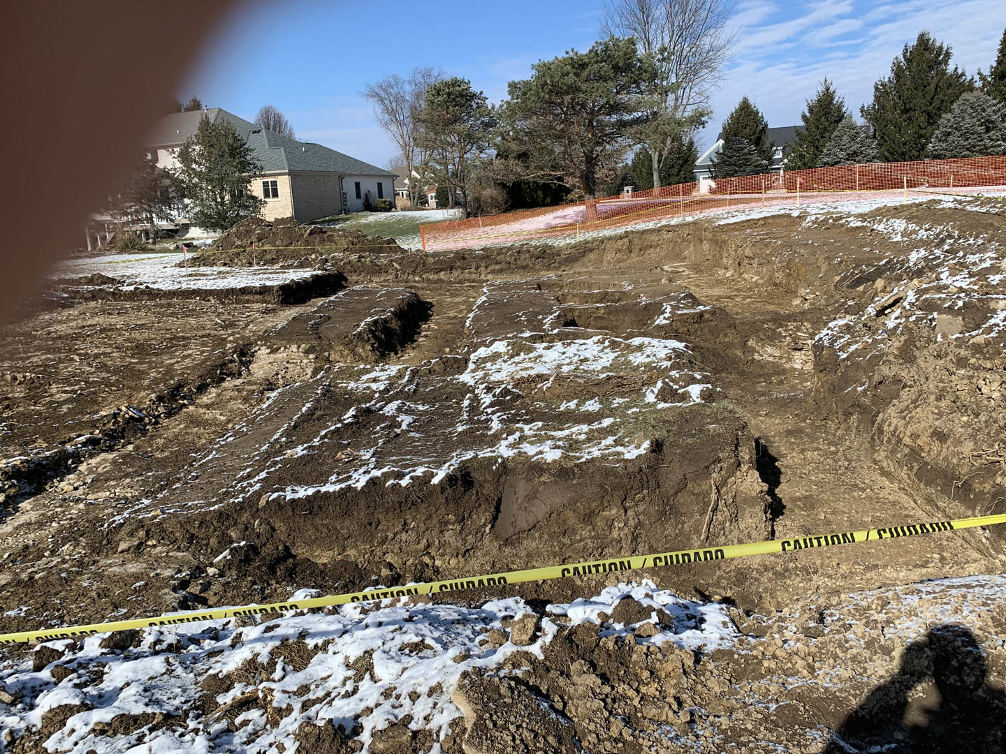 Dirt field with patches of snow, yellow caution tape stretched across a mound, leafless tree and fenced house in the background under a cloudy winter sky