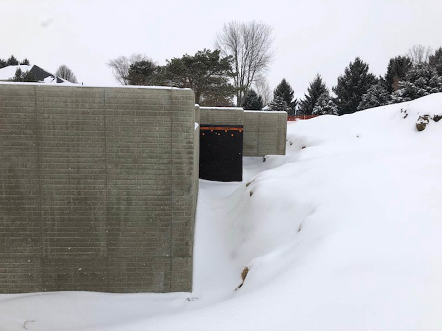 Concrete exterior wall with two black doors, surrounded by snow and leafless trees in winter, white pole and black box with orange stripe visible nearby