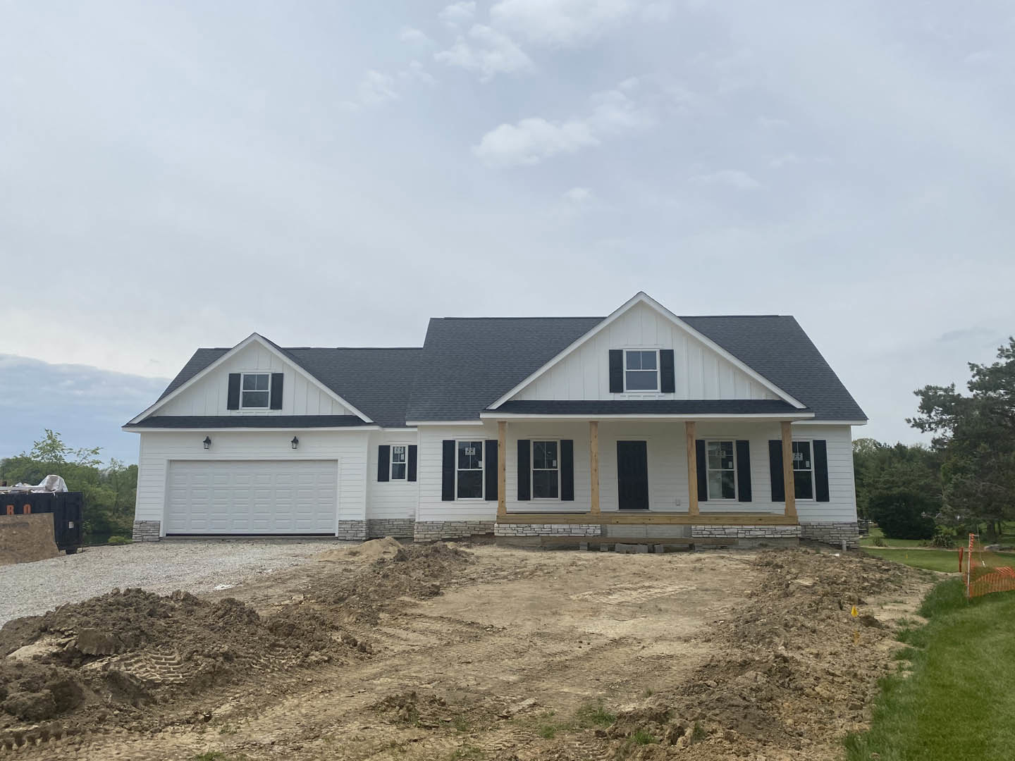 Two-story house under construction with white-framed windows, covered porch, gray roof, surrounded by dirt and patches of grass under a cloudy sky