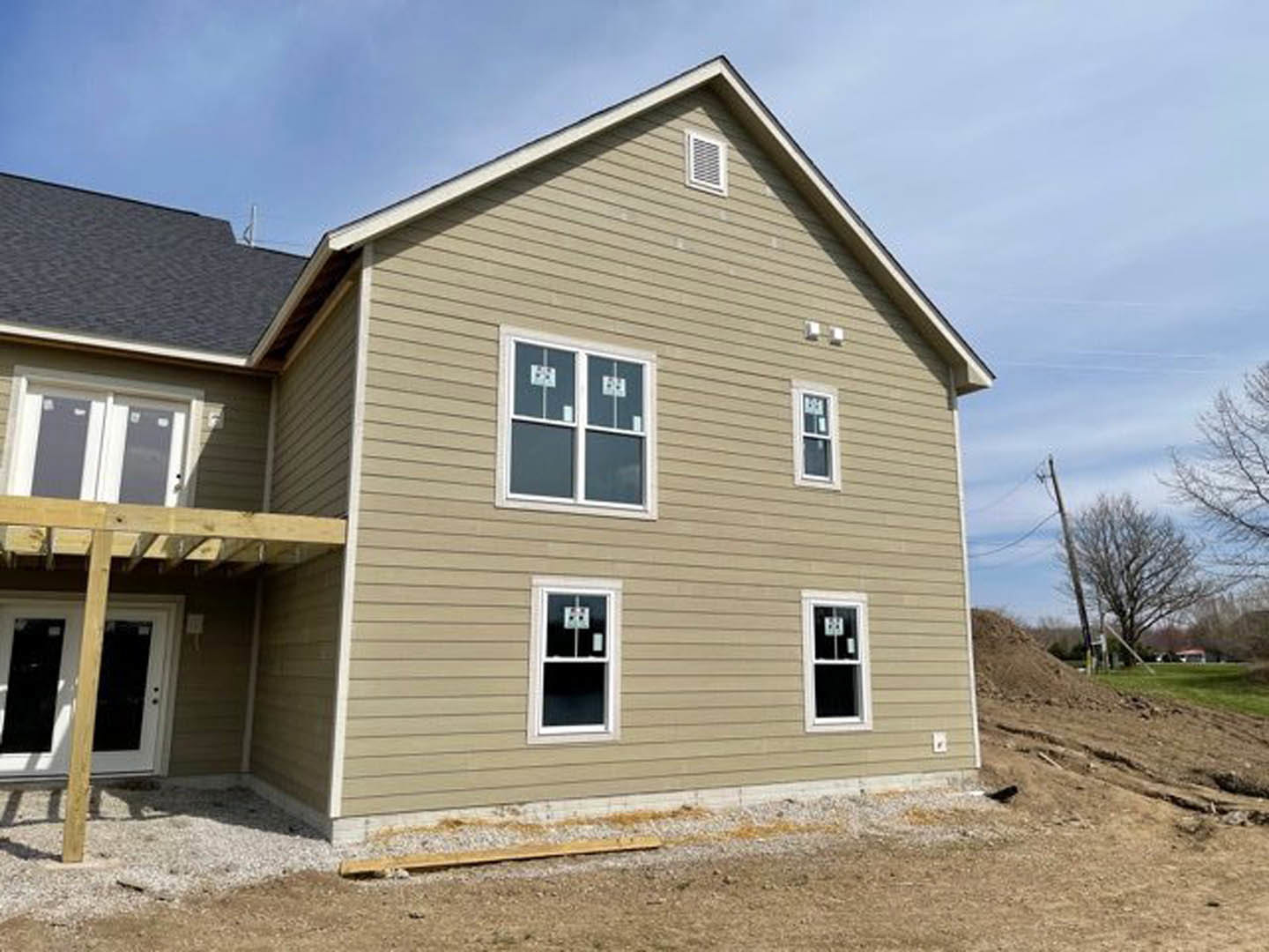 Two-story house under construction with exposed wooden framing, white front door, unfinished porch, dirt mound in foreground, and clear blue sky overhead