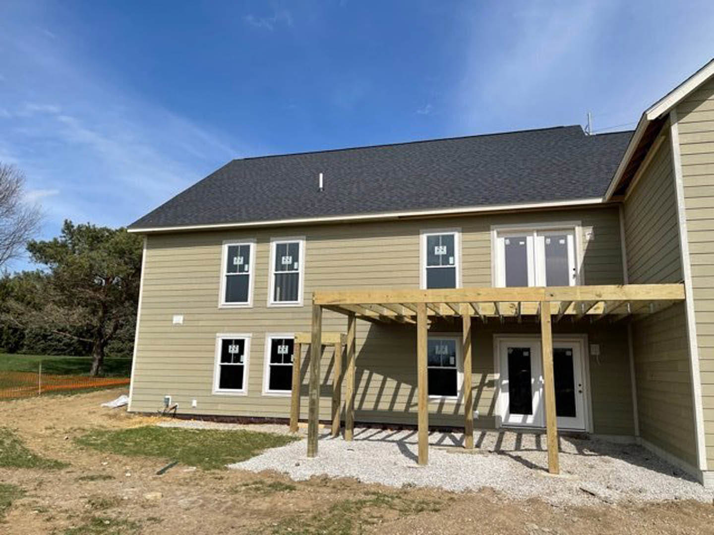 Covered front porch with wooden pergola, white door featuring black glass panels, gray siding, large windows, and landscaped yard with mature tree.