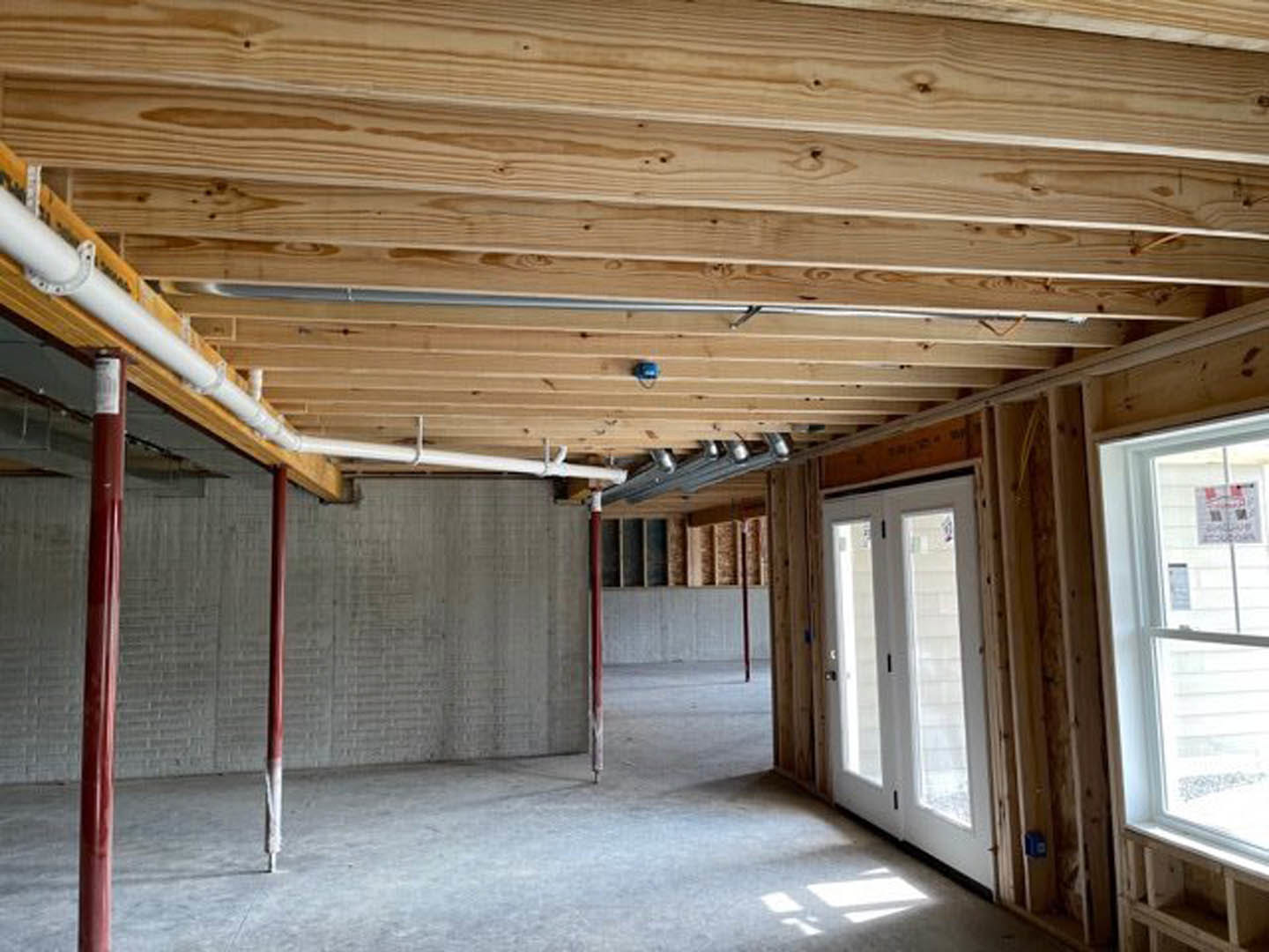Room with exposed wood plank ceiling, visible ceiling pipes, double doors, large window, and contrasting red and black support poles.