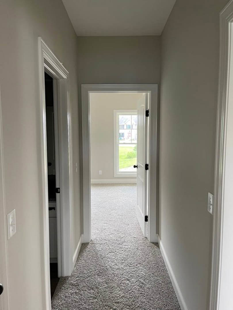 Hallway with white walls and light carpet, open door leading to a room, window overlooking green lawn, visible baseboard molding and plaster finishes