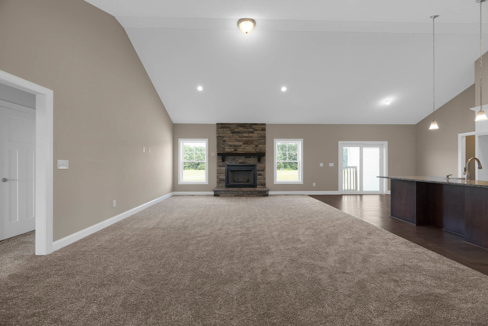 Living room with wood flooring, central fireplace with white mantel, large window showing trees outside, ceiling light fixture, white door with black handle, neutral walls with