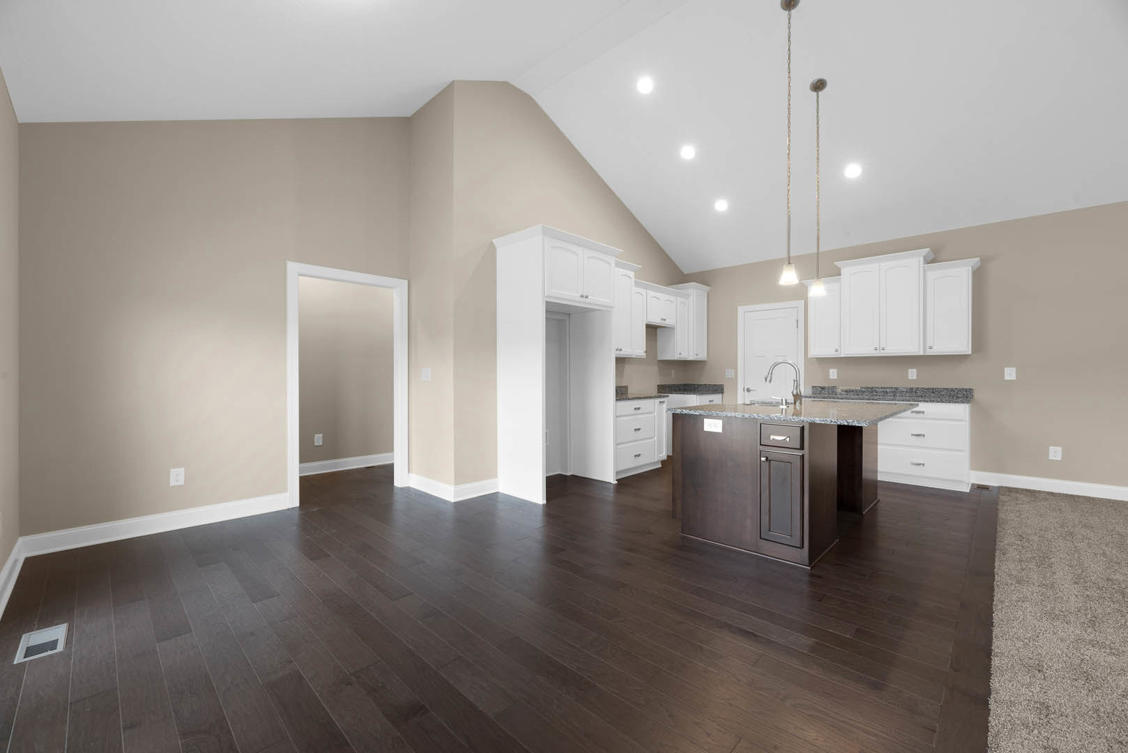 Open kitchen and dining area with dark wood flooring, white walls, white door and frame, vent on wall, carpeted section, and close-up of light-colored kitchen countertop.
