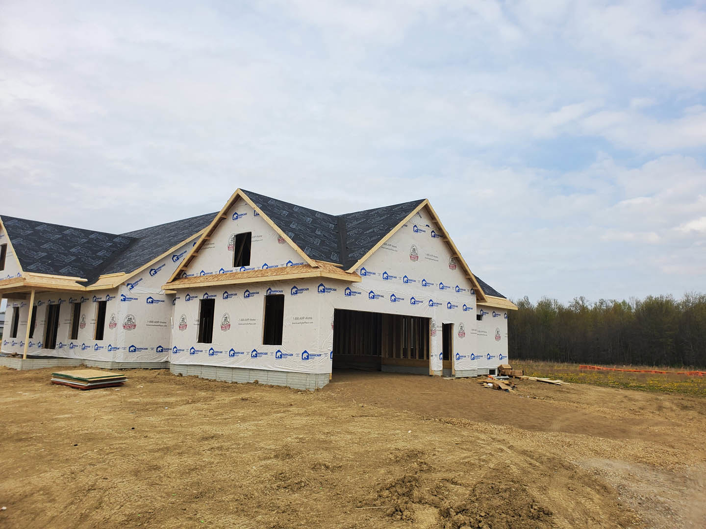 Partially built house with black roof, exposed framing, surrounded by dirt field and mature trees under cloudy sky