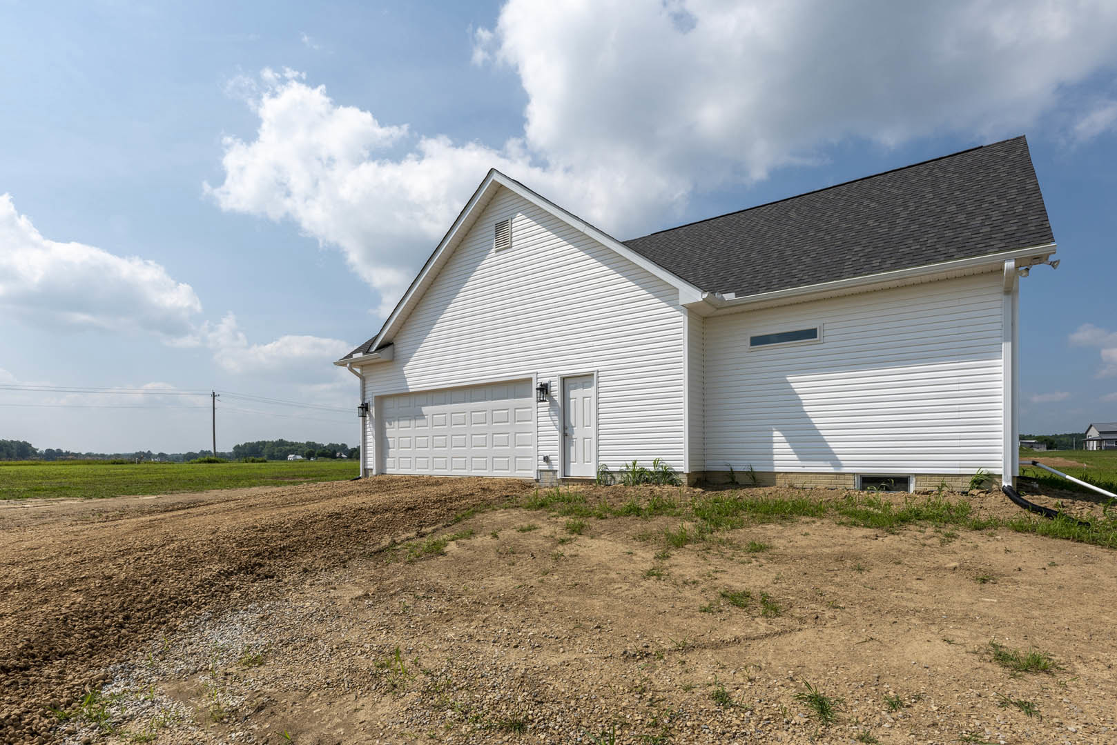 White siding house with attached garage, dirt lot in front, blue sky overhead, sparse grass along property edge