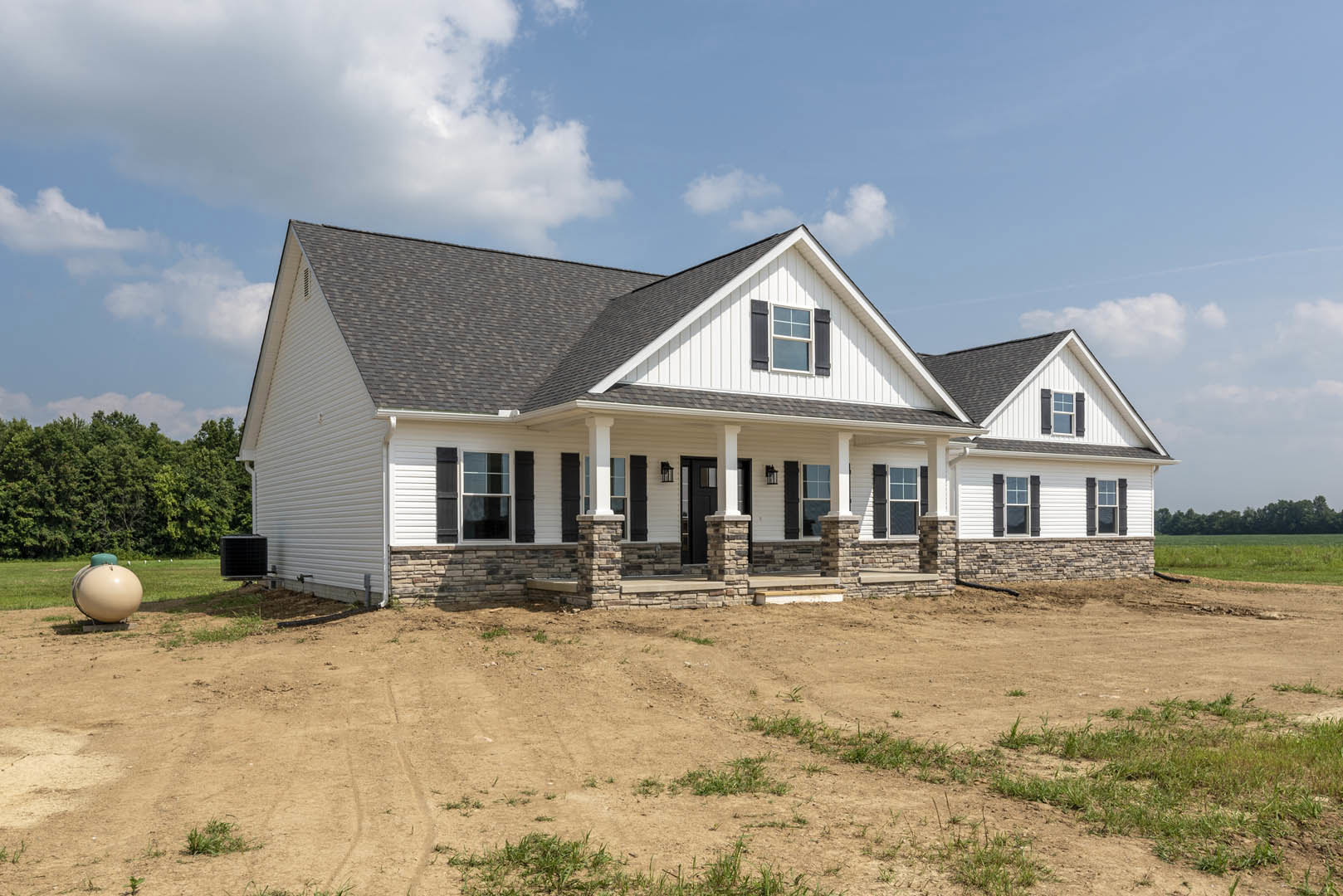 Stone and brick exterior house with covered porch, dirt yard, patchy grass, surrounding trees, and a black mailbox on a stand