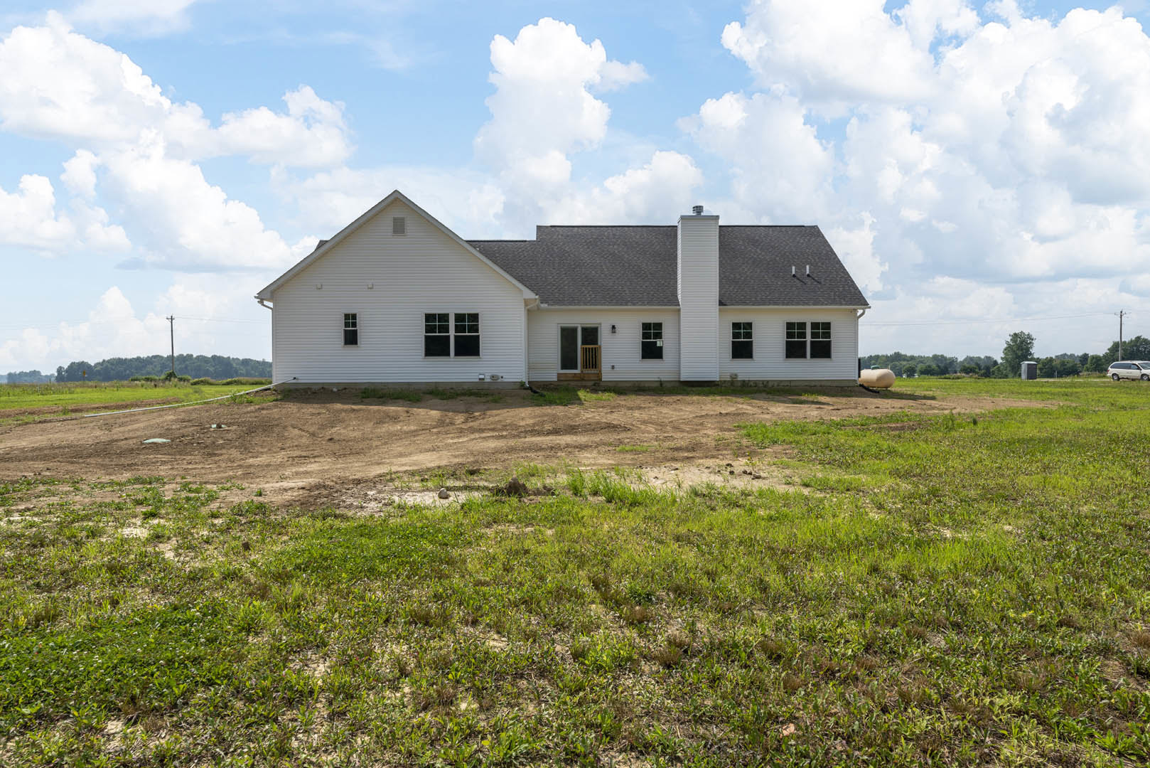 White farmhouse with black roof and covered porch, set on a grassy field under a blue sky with scattered clouds
