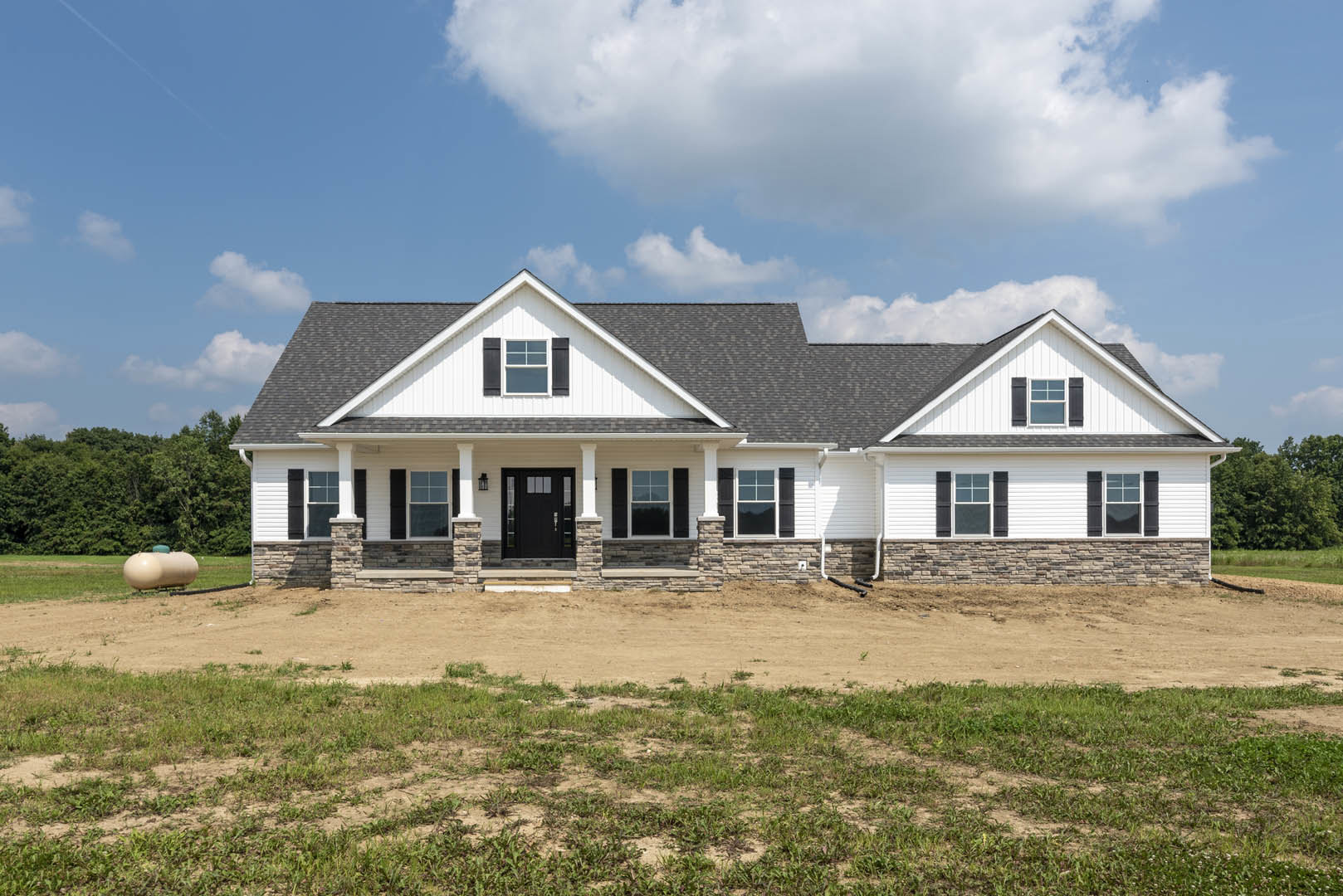 Two-story house with stone pillars, black door with glass panes, surrounded by dirt field and patches of grass under cloudy sky