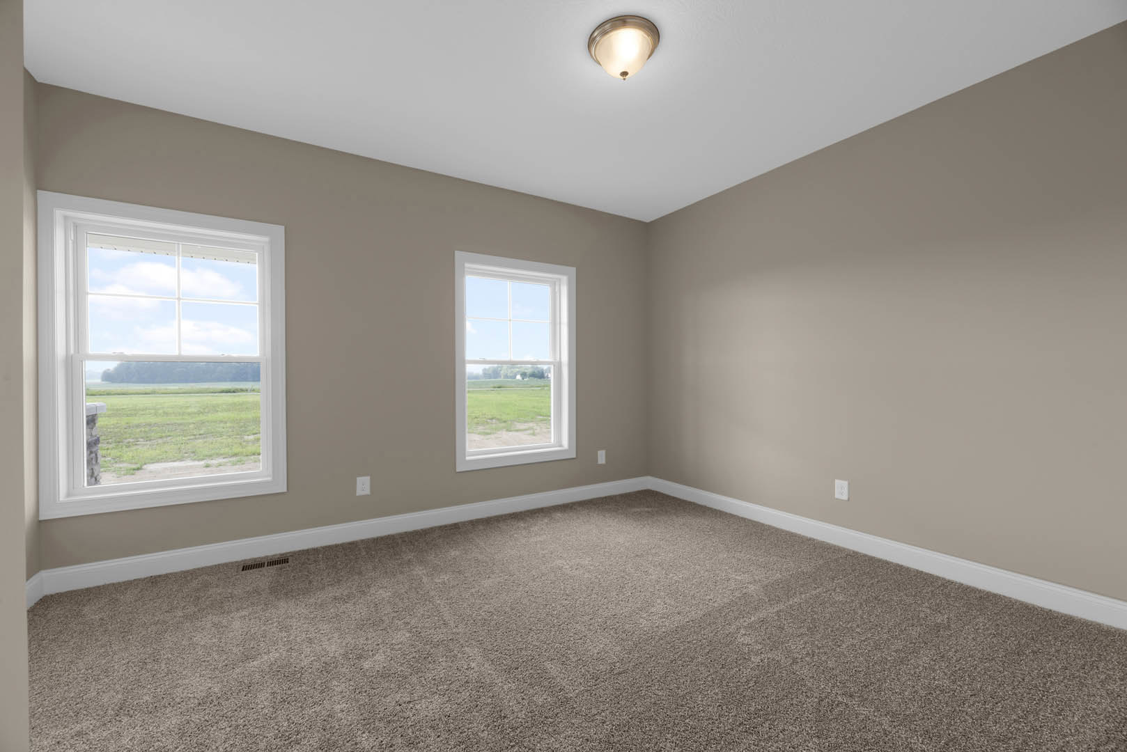 Spacious carpeted room with large windows overlooking a green field and road, white walls, ceiling light fixture, and decorative molding.