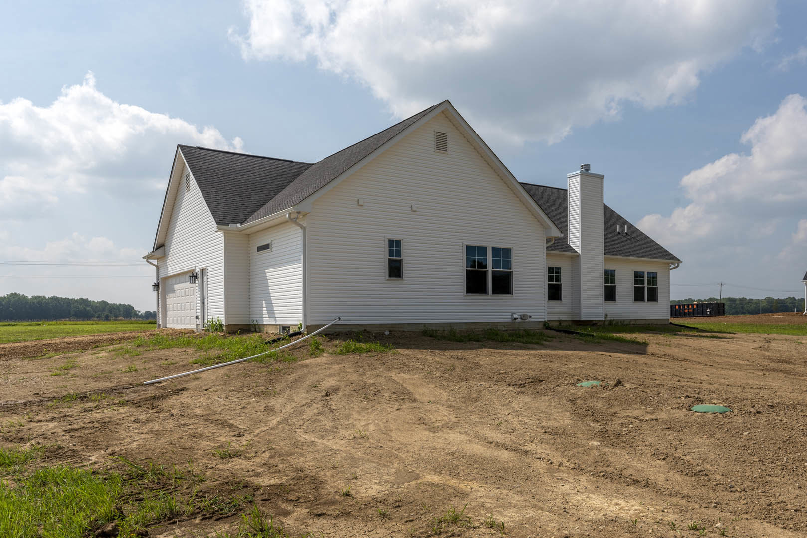 White house with multi-pane windows and white frames, gray roof, dirt patch in foreground with garden hose attached to siding, grassy area and group of trees in the distance under