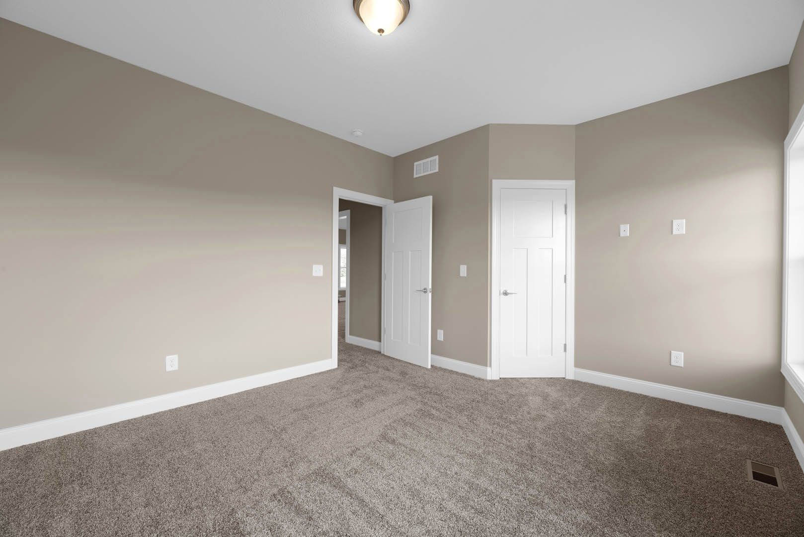 Beige walls and carpeted floor, white door with silver handle, ceiling light fixture, white wall vent, simple molding along baseboards
