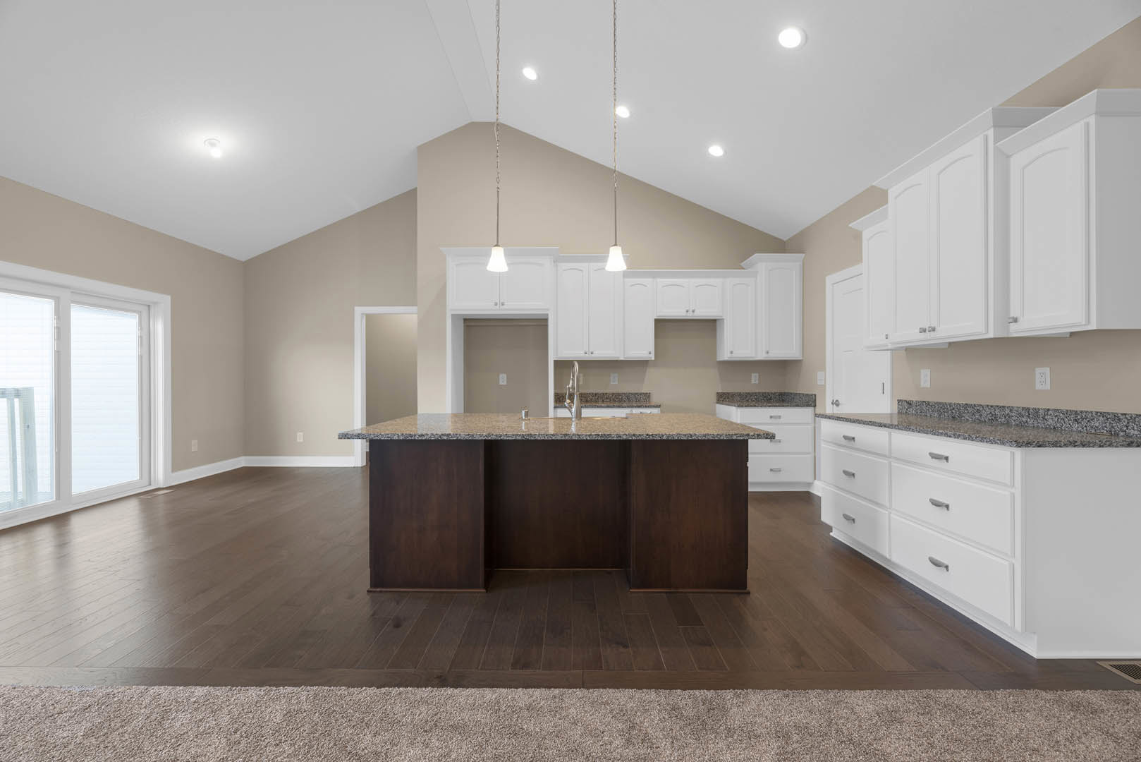 Spacious kitchen featuring a large granite island with wood base, white cabinetry, stainless steel sink, and wide window with white frame; wood and carpet flooring visible.
