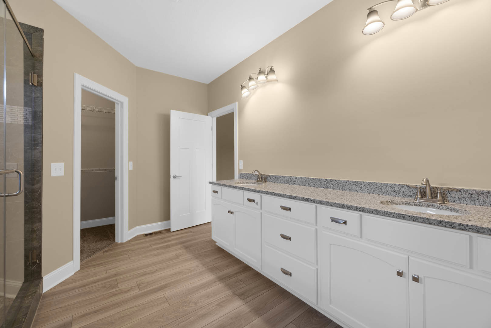 Bathroom with white shaker cabinets, wood plank flooring, white framed door, silver hardware, glass shower stall, and modern light fixture