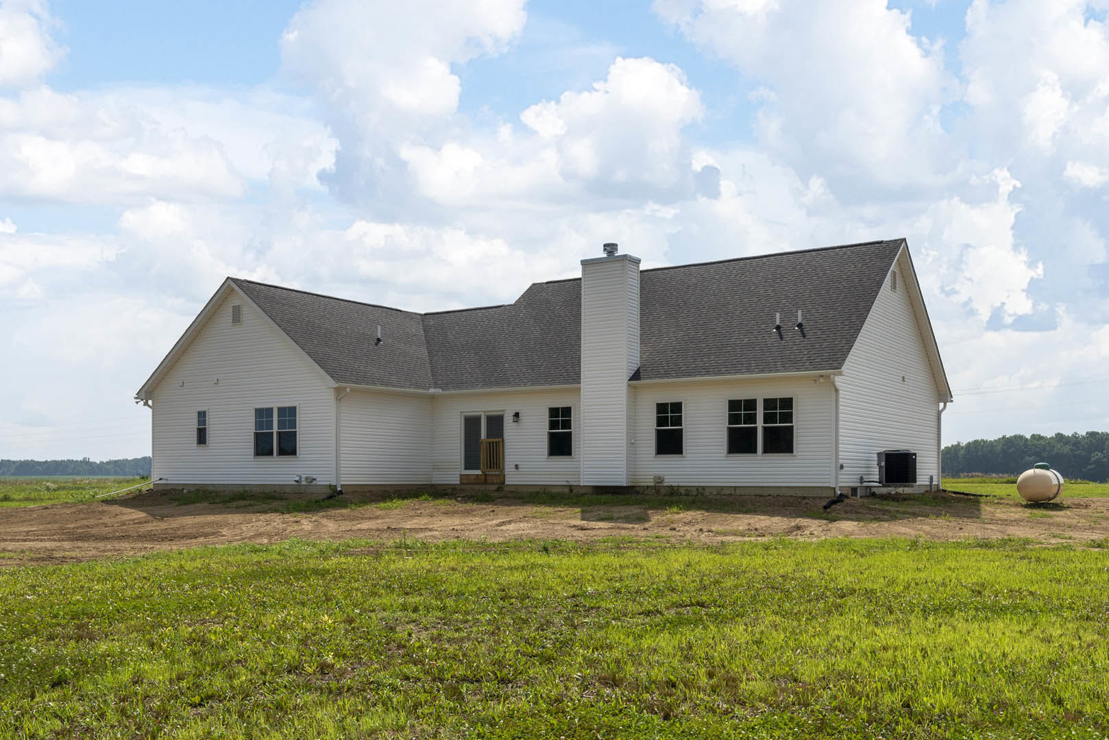 White farmhouse with black roof and chimney, surrounded by grassy field under cloudy sky, windows visible on exterior walls