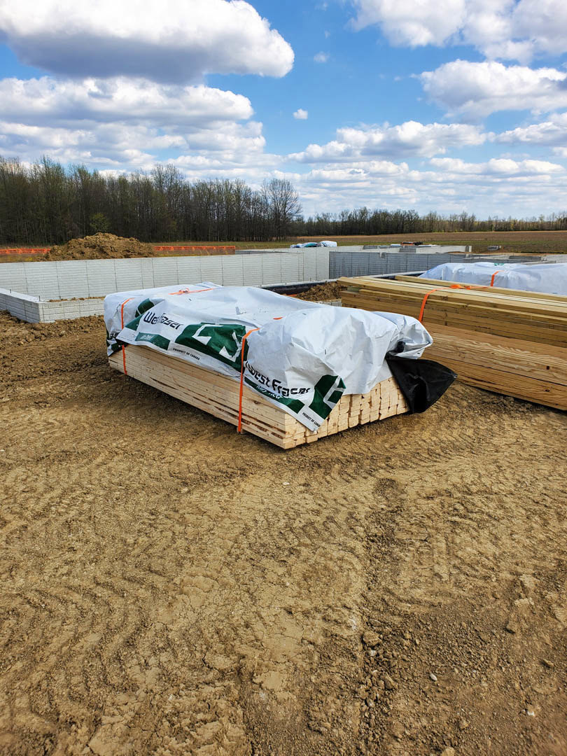 Stacked lumber and wooden pallets covered with a white tarp on a dirt field, surrounded by grass and trees under a blue sky with scattered clouds.