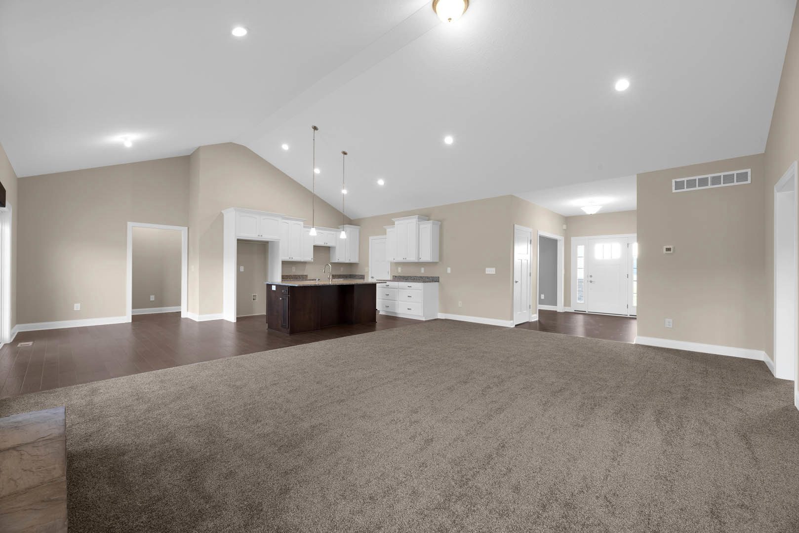 Spacious open floor plan featuring a central kitchen island with white cabinetry, carpeted living area, white door with glass panels, white trim, wall vent, and wood accents