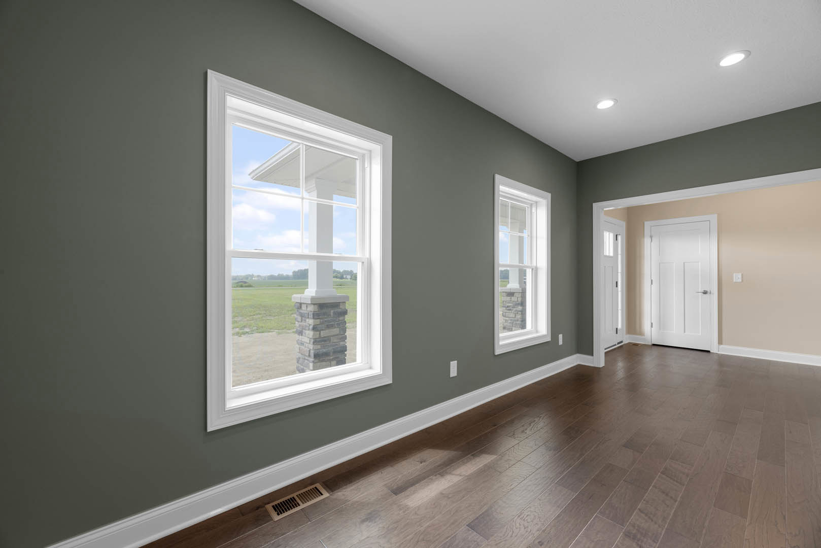 Bright room featuring large white-framed windows, wood flooring with a floor vent, white door with silver handle, and a brick pillar adjacent to the window.
