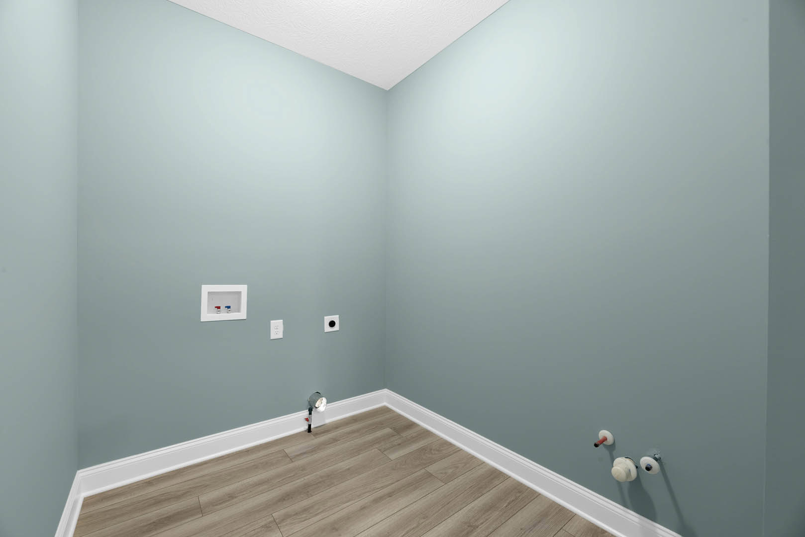 Light blue walls, wood laminate flooring, white ceiling, white wall-mounted box with red and blue faucets, small floor light, white object attached to wall