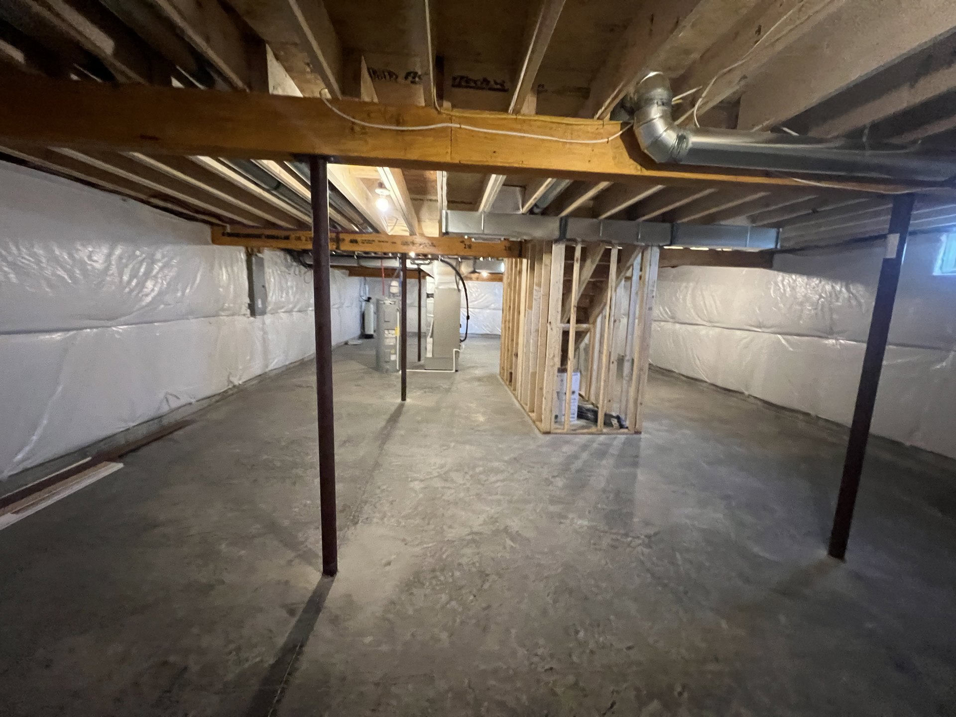 Basement with exposed wooden beam, visible metal pipes, white rectangular object with metal frame, metal support post, grey concrete floor, and white electrical wires along