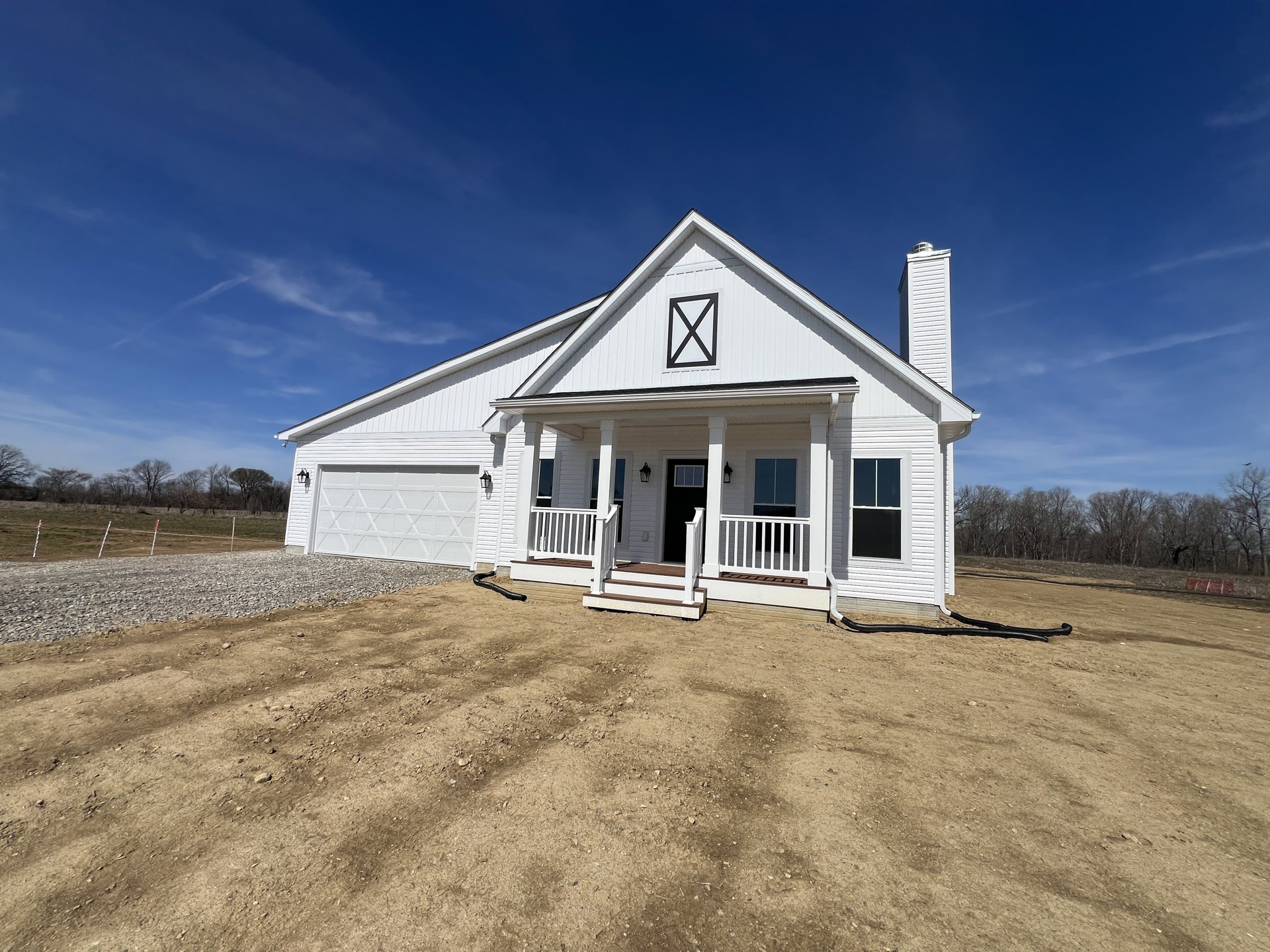 White siding house with attached garage, black square vent on roof, dirt yard, white porch and stairs, white framed windows.