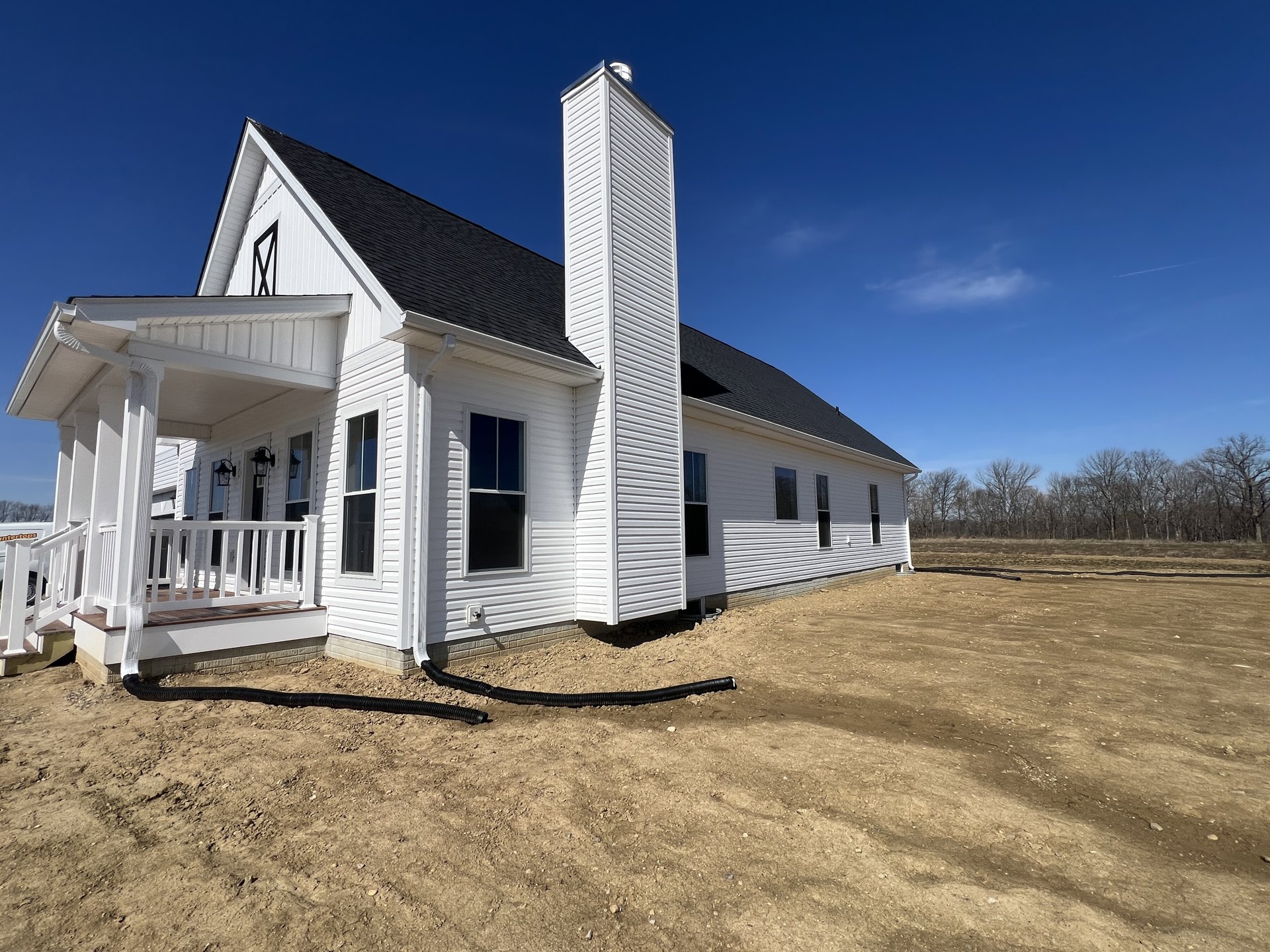 White house with black-framed windows, prominent chimney, covered porch, black pipe on lawn, surrounded by trees under blue sky
